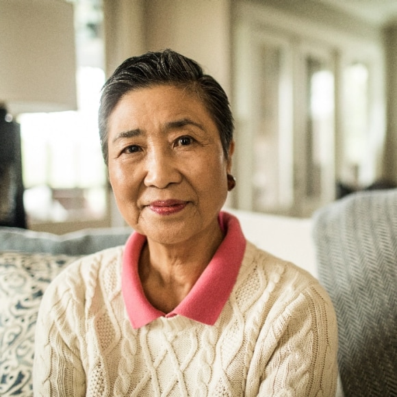 An Asian woman smiles with subtle confidence as she sits on a couch in her home or at a nice bed and breakfast