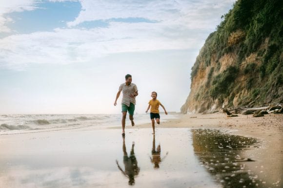 A father and young daughter are all smiles as they run together on a secluded beach