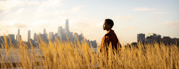 An elegant Black woman walks through wheat grass at sunset with a cityscape in the background