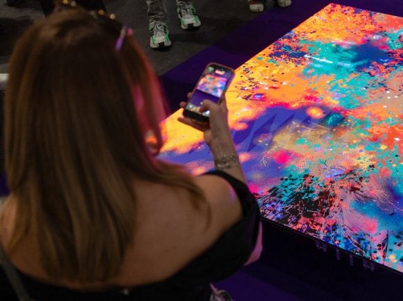 A woman with long light-brown hair, seen from behind, raises her smartphone to capture a photo of a large, flat digital art installation on the floor. The display is filled with a vibrant, chaotic splash of abstract colors of neon orange, hot pink, deep purple, and bright cyan.