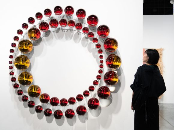 A visitor wearing a black hooded jacket viewing artwork featuring concentric rings of varying-sized glass spheres, painted in gradients of purple, red, and yellow, and mounted on a stainless steel structure 