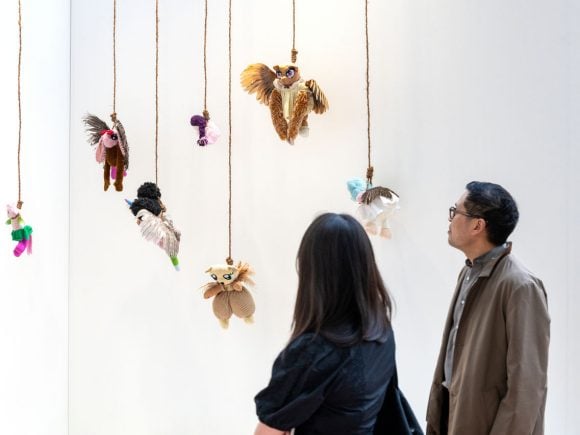 Two viewers observing an art installation featuring  suspended figures and creatures made from discarded children's toys and taxidermized bird wings, hanging from the ceiling