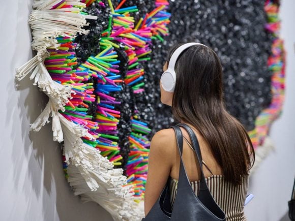 Person wearing white headphones while looking at a large, textured wall sculpture made of colorful, repurposed plastic materials