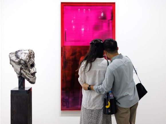 Two visitors observing a large, vivid pink and red gradient monochromatic mirror artwork on a white gallery wall alongside a sculptural piece on a black pedestal