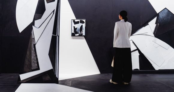 A visitor with dark hair, seen from behind, stands on the black floor wearing a tailored white blazer and wide-leg black trousers, looking at large-scale geometric installation in stark black and white.
