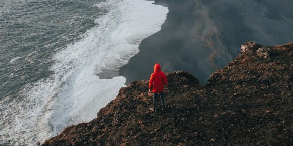 Aerial view of a figure in a red coat standing on a cliff overlooking waves washing up on a dark sandy beach.